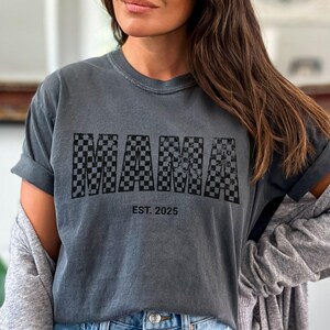 Pu&ograve; includere: T-shirt grigio scuro con la scritta "MAMA" in un motivo a scacchi bianco e nero. Il testo "EST. 2025" &egrave; stampato sotto. La maglietta ha maniche corte e una vestibilit&agrave; rilassata.