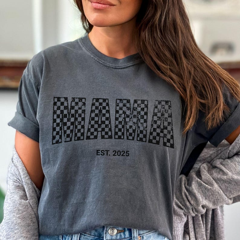 Pu&ograve; includere: T-shirt grigio scuro con la scritta "MAMA" in un motivo a scacchi bianco e nero. Il testo "EST. 2025" &egrave; stampato sotto. La maglietta ha maniche corte e una vestibilit&agrave; rilassata.