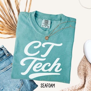 May include: A seafoam green short-sleeve t-shirt with the white text "CT Tech" printed in a cursive font on the front. The shirt features a crew neckline and a Comfort Colors brand label on the inside collar. The word "SEAFOAM" is printed below.
