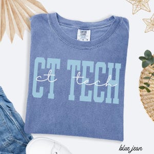 May include: A blue jean colored t-shirt with the words "CT TECH" printed on the front in light blue block letters with the word "tech" in a cursive font. The shirt is made of a soft material and has a crew neck.