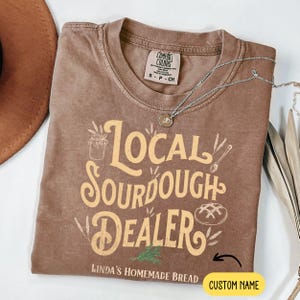 Peut inclure: Un t-shirt marron avec le texte "Local Sourdough Dealer" en lettres jaunes. Le texte "Linda's Homemade Bread" est sous le texte principal, et il y a un espace pour un nom personnalis&eacute; en dessous.