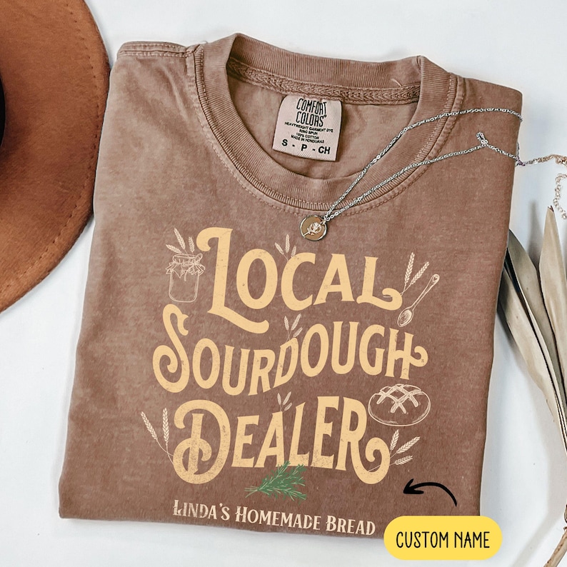 Peut inclure: Un t-shirt marron avec le texte "Local Sourdough Dealer" en lettres jaunes. Le texte "Linda's Homemade Bread" est sous le texte principal, et il y a un espace pour un nom personnalis&eacute; en dessous.