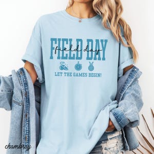 May include: Light blue t-shirt with the words "FIELD DAY" in a bold font, with "field day" in a script font. Below the text are three sports-themed icons and the phrase "LET THE GAMES BEGIN!"