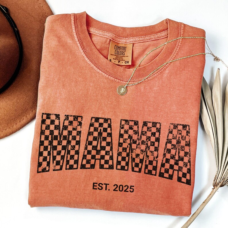 Pu&ograve; includere: T-shirt color corallo con la scritta "MAMA" in un motivo a scacchi neri. Il testo "EST. 2025" &egrave; sotto la parola "MAMA". Sono visibili anche una collana d'oro con un ciondolo e un cappello marrone.