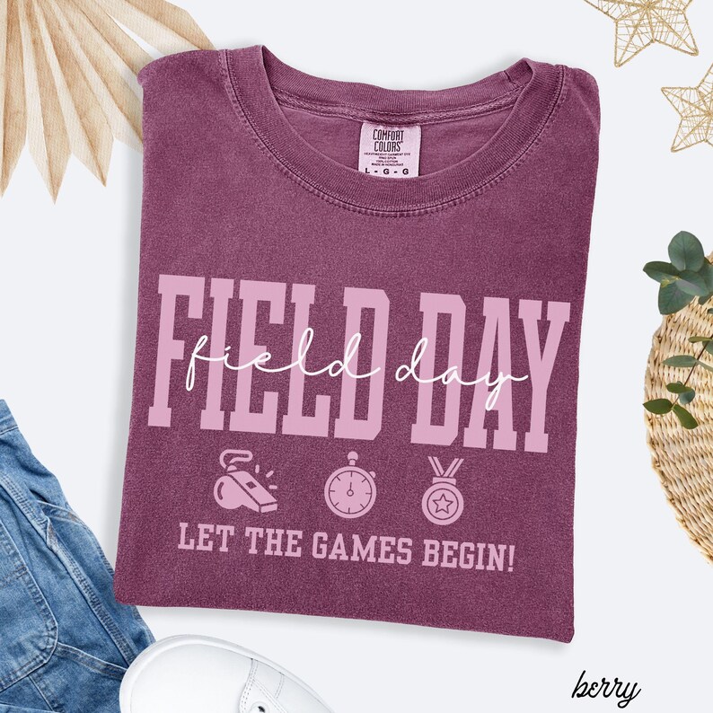 May include: Berry-colored t-shirt with the words "FIELD DAY" in a large, pink font. Below the text are illustrations of a whistle, stopwatch, and medal. The phrase "LET THE GAMES BEGIN!" is printed below the illustrations.
