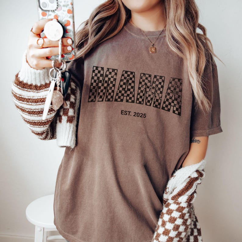 Pu&ograve; includere: T-shirt marrone con la scritta "MAMA" in un motivo a scacchi bianco e nero, con "EST. 2025" stampato sotto. La maglietta &egrave; di colore neutro e ha maniche corte.