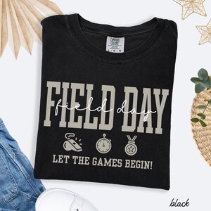 May include: Black t-shirt with the words "FIELD DAY" in large, block letters. The text "field day" is in a cursive font. Below the text are illustrations of a whistle, stopwatch, and medal. The phrase "LET THE GAMES BEGIN!" is printed below the illustrations.