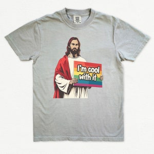 May include: A light gray t-shirt with a cartoon image of Jesus holding a sign that says "I'm cool with it" with a rainbow stripe across the top of the sign.