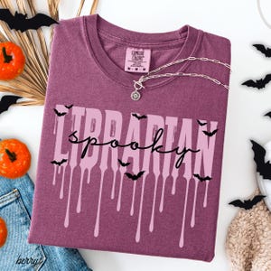 K&ouml;nnte beinhalten: Beerenfarbenes T-Shirt mit den Worten "Librarian Spooky" in einer tropfenden Schriftart, akzentuiert mit schwarzen Fledermaus-Silhouetten. Eine silberne Halskette mit Anh&auml;nger liegt &uuml;ber dem Shirt. Halloween-Dekorationen.