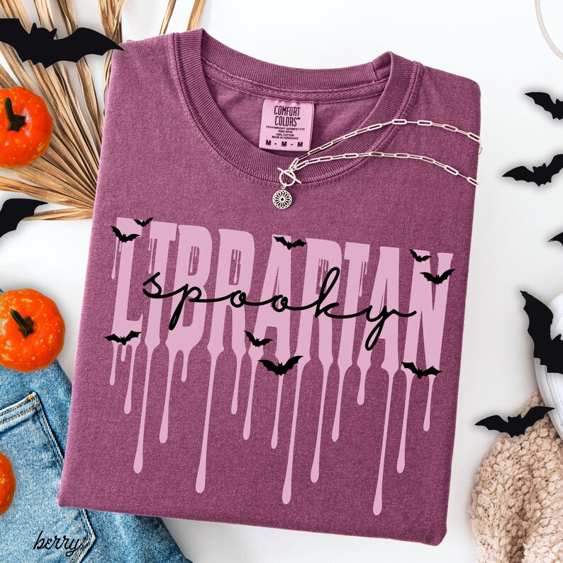 K&ouml;nnte beinhalten: Beerenfarbenes T-Shirt mit den Worten "Librarian Spooky" in einer tropfenden Schriftart, akzentuiert mit schwarzen Fledermaus-Silhouetten. Eine silberne Halskette mit Anh&auml;nger liegt &uuml;ber dem Shirt. Halloween-Dekorationen.