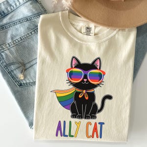 May include: Cream-colored t-shirt featuring a black cat wearing rainbow sunglasses and a rainbow cape. The text "ALLY CAT" is printed below the cat in rainbow colors. The shirt is folded and placed on top of blue jeans and a brown hat.