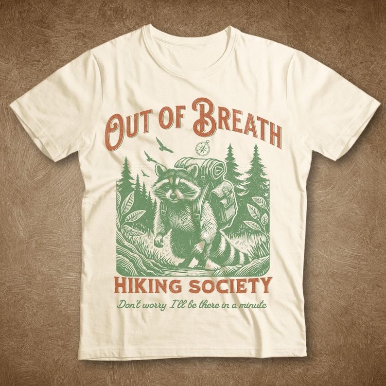 Pode incluir: Uma camiseta branca com uma ilustra&ccedil;&atilde;o verde e marrom de um guaxinim usando uma mochila e caminhando por uma floresta. O texto "Out of Breath Hiking Society" &eacute; impresso acima da ilustra&ccedil;&atilde;o, e o texto "Don't worry I'll be there in a minute" &eacute; impresso abaixo da ilustra&ccedil;&atilde;o.
