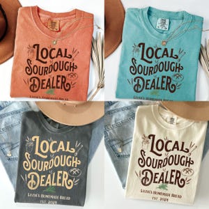 Peut inclure: Quatre t-shirts de couleurs diff&eacute;rentes avec le texte "Local Sourdough Dealer" et "Linda's Homemade Bread" imprim&eacute; dessus. Les t-shirts sont pr&eacute;sent&eacute;s sur une surface en bois avec un jean et un chapeau.