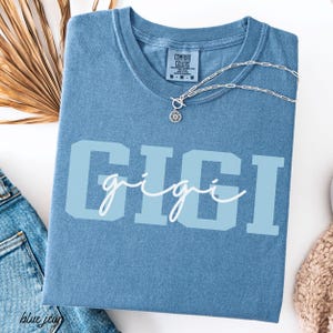 Op de afbeelding: Een blauw Comfort Colors T-shirt met de naam "GIGI" in lichtblauwe blokletters en een cursief lettertype. Een zilveren ketting met een zonnebloemhanger ligt op het shirt. Het shirt is op een wit oppervlak gevouwen.