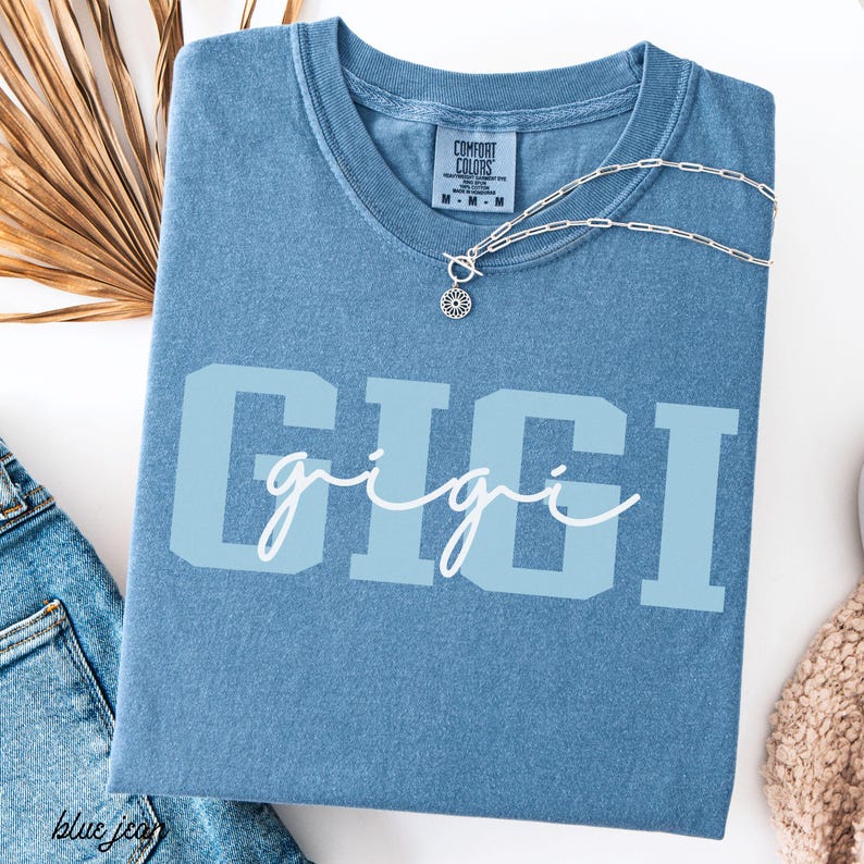 Op de afbeelding: Een blauw Comfort Colors T-shirt met de naam "GIGI" in lichtblauwe blokletters en een cursief lettertype. Een zilveren ketting met een zonnebloemhanger ligt op het shirt. Het shirt is op een wit oppervlak gevouwen.