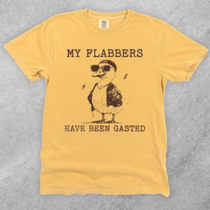 Puede incluir: Una camiseta amarilla con un pato de dibujos animados que lleva gafas de sol y una chaqueta de cuero. El texto "MY FLABBERS HAVE BEEN GASTED" est&aacute; impreso en la camiseta.