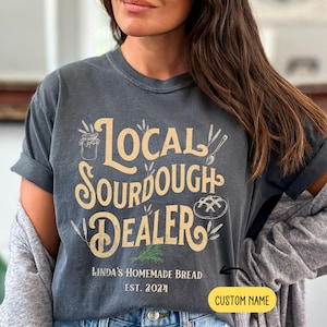 Peut inclure: Un t-shirt gris avec le texte "Local Sourdough Dealer" en lettres jaunes et brunes. Le texte est entour&eacute; d'illustrations d'&eacute;pis de bl&eacute;, d'un pot de levain et d'un pain. Sous le texte se trouve le texte "Linda's Homemade Bread Est. 2024" en lettres noires. Le texte "CUSTOM NAME" est dans une bo&icirc;te jaune dans le coin inf&eacute;rieur droit de l'image.