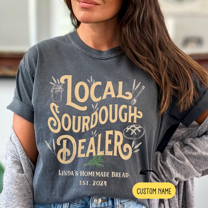 Peut inclure: Un t-shirt gris avec le texte "Local Sourdough Dealer" en lettres jaunes et brunes. Le texte est entour&eacute; d'illustrations d'&eacute;pis de bl&eacute;, d'un pot de levain et d'un pain. Sous le texte se trouve le texte "Linda's Homemade Bread Est. 2024" en lettres noires. Le texte "CUSTOM NAME" est dans une bo&icirc;te jaune dans le coin inf&eacute;rieur droit de l'image.