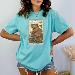 Pode incluir: Uma camiseta azul turquesa com um gr&aacute;fico de uma carta de tar&ocirc; que apresenta uma capivara em um campo de flores. A carta &eacute; etiquetada como "XIX O Capybara".