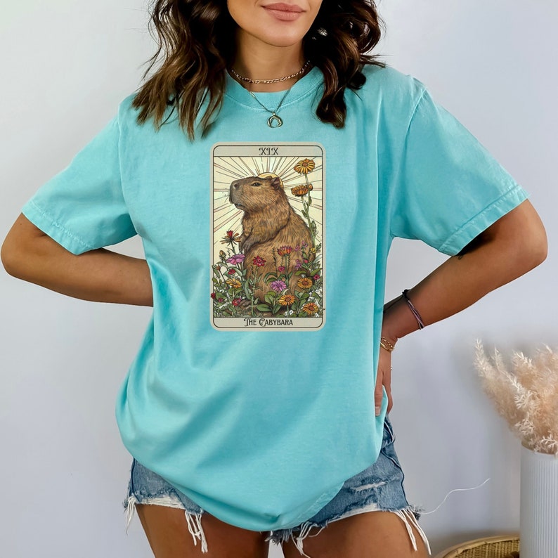 Pode incluir: Uma camiseta azul turquesa com um gr&aacute;fico de uma carta de tar&ocirc; que apresenta uma capivara em um campo de flores. A carta &eacute; etiquetada como "XIX O Capybara".