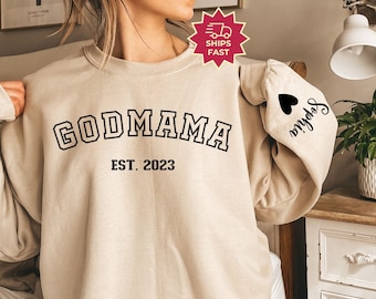 May include: A cream-coloured sweatshirt displaying the text "GODMAMA" in black, with "EST. 2023" underneath. The sleeve has a black heart and the name "Sophia" in script. A "SHIPS FAST" graphic is also visible.