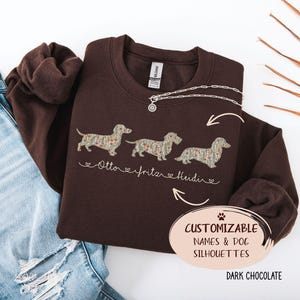 Könnte beinhalten: Ein dunkel schokoladenbraunes Sweatshirt mit Rundhalsausschnitt und drei gestickten Dackel-Silhouetten mit Blumenmuster. Unter den Hunden sind die Namen Otto, Fritz und Heidi in weißer Schreibschrift gestickt. Ein Etikett zeigt CUSTOMIZABLE NAMES & DOG SILHOUETTES.