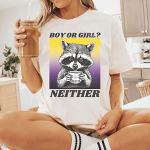 Pode incluir: Camiseta branca com um gráfico de um guaxinim segurando uma xícara de café. O texto "Boy or Girl? Neither" está impresso na camiseta.