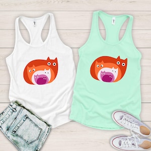 May include: Two tank tops, one white and one mint green, each featuring a graphic of stylized cats in shades of orange, pink, and white. The white tank top is paired with denim shorts, while the green one is next to white sneakers.