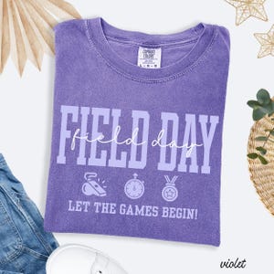 May include: Violet Comfort Colors t-shirt with the words "FIELD DAY" in large white letters. Below the text are three sports-themed icons and the phrase "LET THE GAMES BEGIN!". The shirt is folded and lies on a white surface.