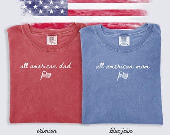 Pode incluir: Duas camisetas Comfort Colors, uma carmesim e outra azul jeans, com o texto "all american dad" e "all american mom" em escrita branca, junto com um pequeno gráfico de bandeira. Um gráfico da bandeira americana no fundo.