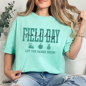 May include: A light green t-shirt with the words "FIELD DAY" in dark green block letters and a cursive "day" in the middle. Below the text are three sports-themed icons and the phrase "LET THE GAMES BEGIN!"