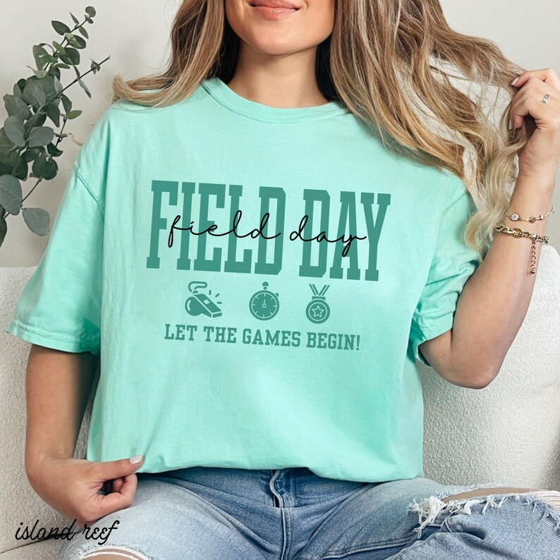 May include: A light green t-shirt with the words "FIELD DAY" in dark green block letters and a cursive "day" in the middle. Below the text are three sports-themed icons and the phrase "LET THE GAMES BEGIN!"