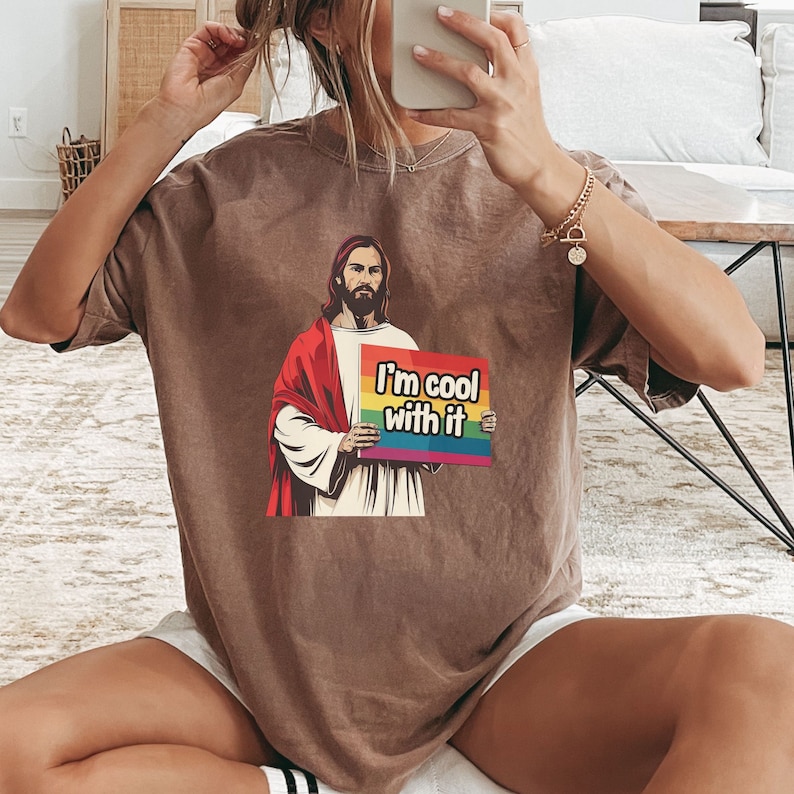 May include: A brown t-shirt with a graphic of Jesus holding a sign that says "I'm cool with it". The sign has a rainbow stripe across the top.