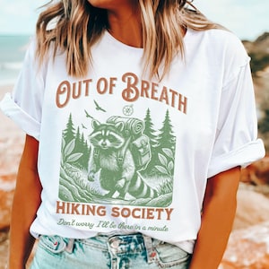 Pode incluir: Camiseta branca com um guaxinim verde usando uma mochila e uma b&uacute;ssola. O texto "Out of Breath Hiking Society" est&aacute; impresso na camiseta. O texto "Don't worry I'll be there in a minute" est&aacute; impresso abaixo do gr&aacute;fico.