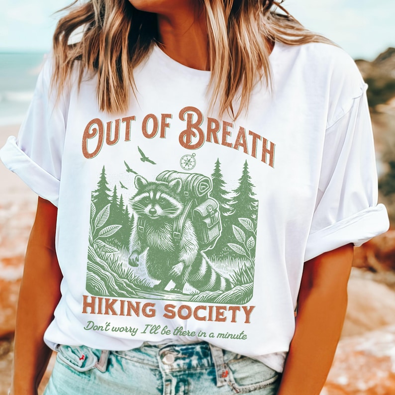Pode incluir: Camiseta branca com um guaxinim verde usando uma mochila e uma b&uacute;ssola. O texto "Out of Breath Hiking Society" est&aacute; impresso na camiseta. O texto "Don't worry I'll be there in a minute" est&aacute; impresso abaixo do gr&aacute;fico.