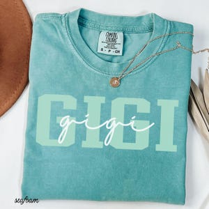 Op de afbeelding: Zeegroen t-shirt met het woord "GIGI" in grote, lichtgroene blokletters en een cursieve "gigi" eronder. Een gouden ketting met een kleine hanger ligt op het shirt. Het shirt heeft een Comfort Colors label.