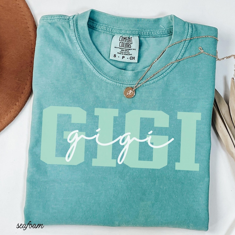 Op de afbeelding: Zeegroen t-shirt met het woord "GIGI" in grote, lichtgroene blokletters en een cursieve "gigi" eronder. Een gouden ketting met een kleine hanger ligt op het shirt. Het shirt heeft een Comfort Colors label.