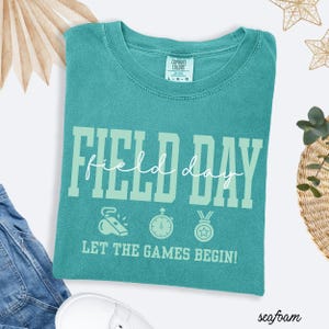 May include: Seafoam green t-shirt with the words "FIELD DAY" in a light green font. Below the text are illustrations of a whistle, stopwatch, and medal. The phrase "LET THE GAMES BEGIN!" is printed below the illustrations.