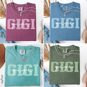 Op de afbeelding: Vier verschillend gekleurde t-shirts, elk met de naam "GIGI" op de voorkant gedrukt. De shirts zijn in bessen, spijkerbroekblauw, zeegroen en mosgroen. Elke shirt heeft een zilveren ketting met een hanger.