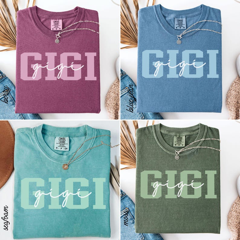 Op de afbeelding: Vier verschillend gekleurde t-shirts, elk met de naam "GIGI" op de voorkant gedrukt. De shirts zijn in bessen, spijkerbroekblauw, zeegroen en mosgroen. Elke shirt heeft een zilveren ketting met een hanger.