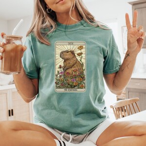 Pode incluir: Uma camiseta azul turquesa com um gr&aacute;fico de uma carta de tar&ocirc; que apresenta uma capivara. A carta &eacute; etiquetada como "XIX O Capivara".