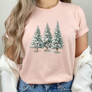 K&ouml;nnte beinhalten: Hellrosa T-Shirt mit einem Aquarelldesign von drei Weihnachtsb&auml;umen. Jeder Baum hat eine rosa Schleife. Die B&auml;ume sind gr&uuml;n mit wei&szlig;en Akzenten. Das T-Shirt ist einfarbig.