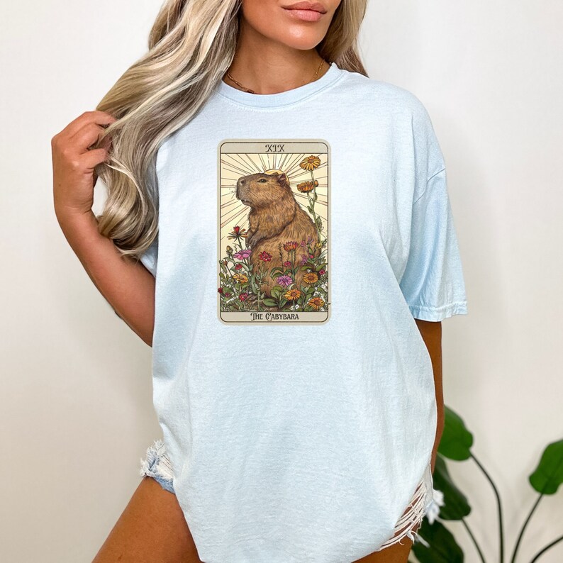 Pode incluir: Uma camiseta azul claro com um gr&aacute;fico de uma carta de tar&ocirc; com uma capivara cercada de flores. A carta &eacute; rotulada como "XIX O Capybara".