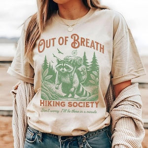 Pode incluir: Uma camiseta bege com um gr&aacute;fico de um guaxinim usando uma mochila e fazendo caminhadas na floresta. O texto "Out of Breath Hiking Society" est&aacute; impresso na camiseta, junto com a frase "Don't worry I'll be there in a minute".