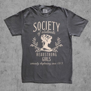 May include: A dark gray t-shirt with a white graphic design. The graphic features a woman's silhouette with a laurel wreath and the text "Society of obstinate Headstrong Girls seriously displeasing since 1813".