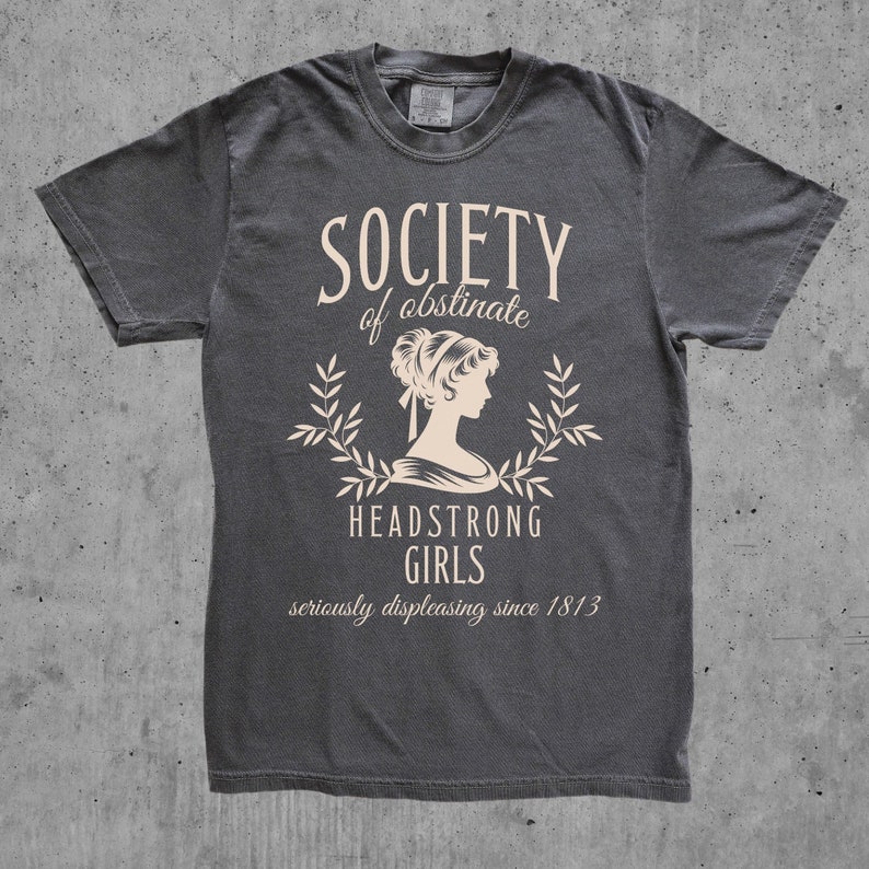 May include: A dark gray t-shirt with a white graphic design. The graphic features a woman's silhouette with a laurel wreath and the text "Society of obstinate Headstrong Girls seriously displeasing since 1813".