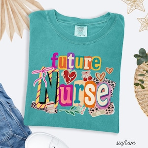 Może przedstawiać: Miętowa koszulka z napisem "future Nurse" w kolorowym, zabawnym wzorze. Grafika zawiera serca, wiśnie i filiżankę kawy. Koszulka jest wykonana z miękkiego materiału.