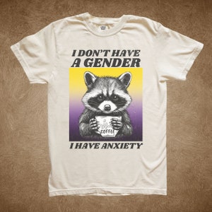 May include: A white t-shirt with a black and white illustration of a raccoon holding a coffee cup. The text "I don't have a gender I have anxiety" is printed on the shirt.