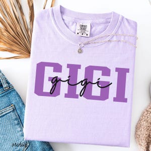 Op de afbeelding: Een lichtpaars Comfort Colors T-shirt met de naam "GIGI" in grote paarse blokletters en een kleiner cursief lettertype. Een delicate zilveren ketting met een zonnebloemhanger ligt op het shirt. Het shirt ligt op een witte ondergrond.