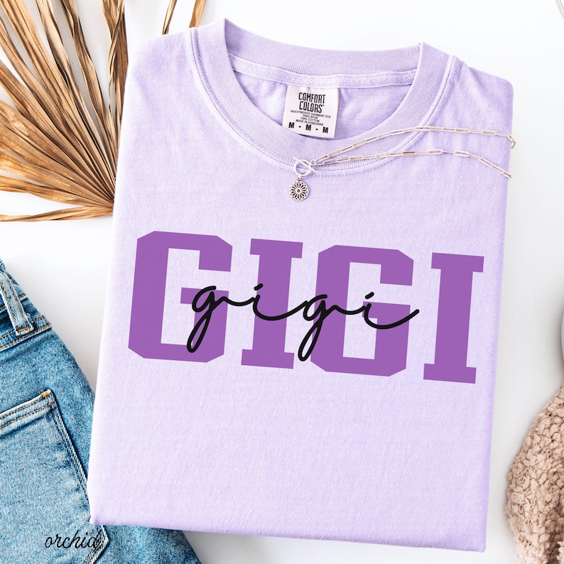 Op de afbeelding: Een lichtpaars Comfort Colors T-shirt met de naam "GIGI" in grote paarse blokletters en een kleiner cursief lettertype. Een delicate zilveren ketting met een zonnebloemhanger ligt op het shirt. Het shirt ligt op een witte ondergrond.